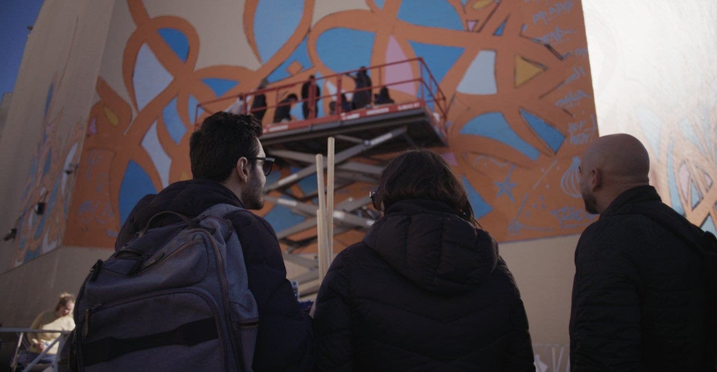 Onlookers watch artists paint a large-scale mural from a scissor lift