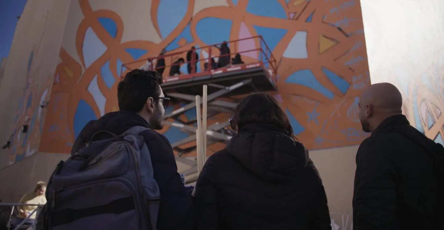 Onlookers watch artists paint a large-scale mural from a scissor lift
